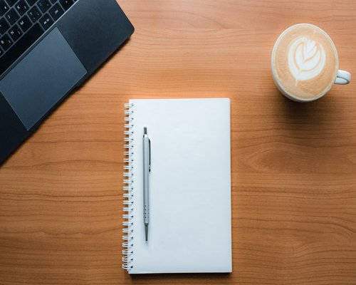 A notebook, pen, and coffee cup on a wooden table setup for work